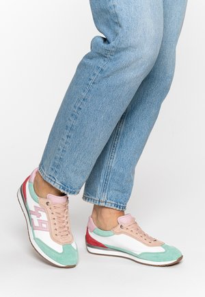 Persona con jeans azzurri e sneakers dai colori pastello con dettagli rosa, verdi, bianchi e rossi, in piedi su una superficie bianca.