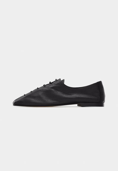 Chaussure plate en cuir noir avec un bout pointu, coutures minimales et détails de boutons bouclés le long du dessus du cou-de-pied.