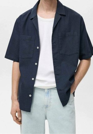 Homme portant une chemise bleu marine à manches courtes boutonnée sur un t-shirt blanc avec un jean bleu clair, une main dans la poche et une à ses côtés.
