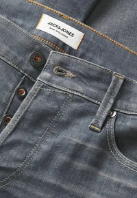 Tamne traperice u tamnoplavoj boji s suptilnom teksturom, s oznakom JACK & JONES, bakrenim zakovicama i kontrastnim šavovima.