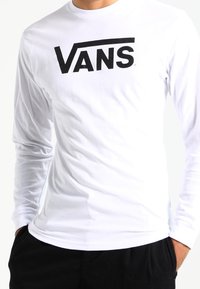 Pitkähihaisen valkoisen t-paidan rinnassa on suuri musta "VANS" -logo. Paidassa on sileä puuvillapinta ja pyöreä kaula-aukko.
