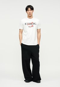 Άνδρας που στέκεται με τα χέρια στις τσέπες, φορώντας λευκό T-shirt με σχέδιο και φαρδιά μαύρα παντελόνια, μπροστά σε απλό φόντο.