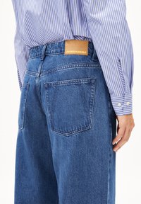 Jeans in denim blu con vita alta, caratterizzati da tasche posteriori, un'etichetta in pelle beige cucita vicino all'orlo della vita e un tessuto testurizzato.