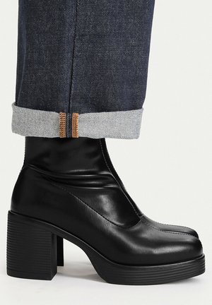 Sort læder ankelstøvle med høje hæle båret med opsmøgede mørkeblå denim-jeans mod en neutral baggrund.