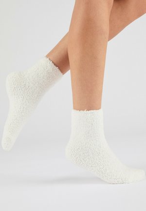 Jambes portant des chaussettes blanches, douces et duveteuses, aux chevilles sur un fond gris clair, un pied légèrement relevé.
