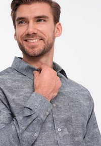 Camisa de botones gris con estampado floral y cuello extendido, con costuras y botones claros, usada por un modelo masculino sonriente.