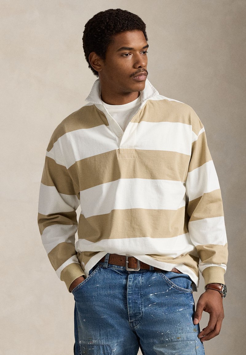 Μακρυμάνικο πουκάμισο ράγκμπι σε μπεζ και λευκές ρίγες, με λευκό γιακά, συνδυασμένο με τζιν παντελόνι και καφέ ζώνη. Φόντο με υφή ξύλου.