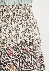 Jupe en coton avec une taille smockée. Présente des motifs floraux noirs et oliviers sur un fond crème, avec des accents géométriques en rouge et noir.
