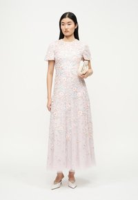 Γυναίκα ντυμένη με ανοιχτό ροζ τούλινο φόρεμα κεντημένο με λουλούδια και κοντά μανίκια, κρατώντας λευκό τσαντάκι και φορώντας λευκά τακούνια.