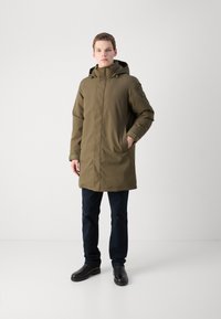 Olivgrüner Parka, knielang, mit Kapuze, vorn mit Reißverschluss und zwei Seitentaschen. Über dunklen Hosen und stabilen schwarzen Schuhen getragen.