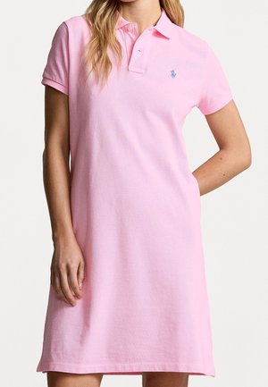 Kobieta ubrana w różową sukienkę polo z krótkim rękawem, kołnierzykiem i plisą na guziki, z małym niebieskim logo na piersiach.