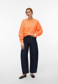 Femme debout portant un pull orange vif, un jean large foncé et des ballerines noires sur fond blanc.