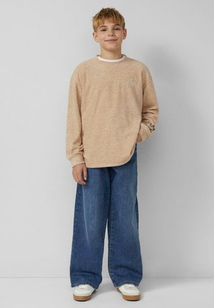 Hellbeiges Sweatshirt mit langen Ärmeln und lockerer Passform, kombiniert mit dunkelblauen Jeans in weiter Beinführung. Weiße Sneaker mit braunen Sohlen.