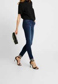 Schmale Jeans aus dunkelblauem Denim mit einer Mittelhoch taille, kombiniert mit schwarzen High-Heelsandaletten. Das Outfit wird durch eine schwarze Clutch ergänzt.