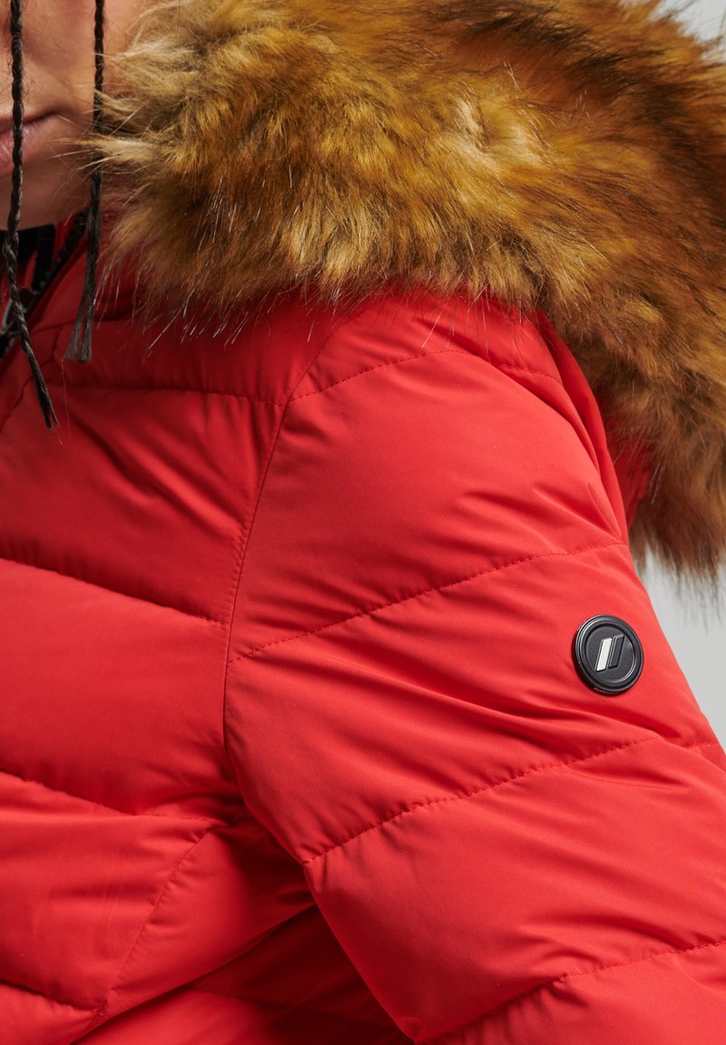 Rode gewatteerde jas met een nep bont capuchon, diagonale gestikte textuur en een zwart rond logo accent op de linkerarm.