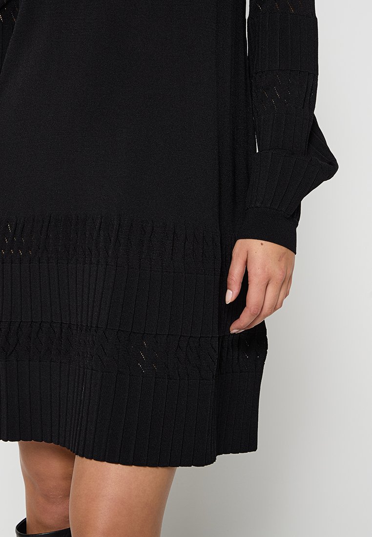 Robe noire tricotée avec une texture côtelée, des motifs ajourés sur les manches et l'ourlet, et une coupe décontractée. Vue rapprochée du tissu et de la main.