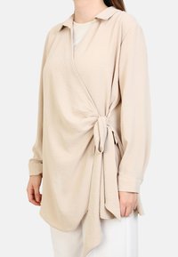 Beige wrap top, joka on valmistettu pehmeästä kankaasta ja jossa on vyösolmu vyötäröllä sekä kärkitakki. Pitkät hihat ja rento istuvuus.