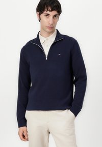 Marineblauwe zip-up trui met geribbelde manchetten en onderrand, voorzien van een klein logo op de borst. Draag over een beige overhemd met kraag.