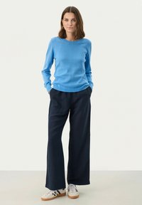 Gai zilas džemperis pieguļošā dizainā, apvienots ar brīvām tumši zilām platu kāju biksēm. Apģērbu papildina baltas sporta kurpes.