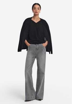 Schwarzer, langärmeliger Cardigan über einem schwarzen V-Ausschnitt-Top, kombiniert mit grauen Jeans mit hohem Bund und weitem Bein. Schlichtes Design mit glatter Textur.