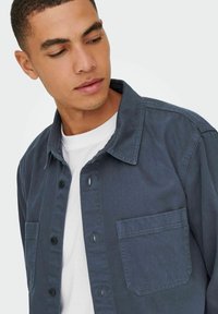 Camicia blu scuro con bottoni, realizzata in tessuto testurizzato, con colletto all'americana, tasche frontali e sottoshirt bianco a contrasto.