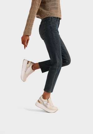 Persona che indossa sneaker beige con accenti dorati, jeans blu scuro e un top a maniche lunghe beige lavorato a maglia, posando con una gamba piegata su sfondo bianco.