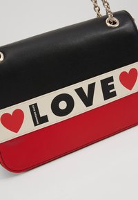 Černá a červená kožená crossbody taška se bílým pruhem s nápisem "LOVE" a grafikou červeného srdce. Řetízkové detaily dodávají funkční akcent.