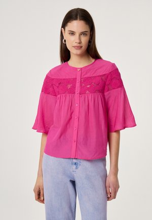 Frau trägt eine leuchtend pinke Bluse mit Knöpfen, Spitzenverzierung über der Brust und ausgestellten Ärmeln, kombiniert mit hellblauen Jeans.