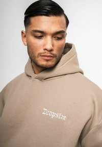 Beige Baumwoll-Hoodie mit einer vorderen Kängurutasche und einem verstellbaren Kordelzug an der Kapuze. Mit weißem gesticktem Text "Dropsize" auf der Brust.