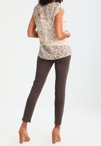 Blusa trasparente con motivo floreale beige, abbinata a pantaloni scuri aderenti marroni e tacchi alti beige. Tessuto liscio con vestibilità comoda.