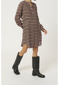 Langärmliges Kleid in geometrischem Muster aus Rosa, Braun und Schwarz; knielang mit einem geknöpften Ausschnitt. Getragen mit kniehohen schwarzen Stiefeln.