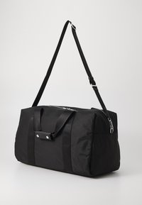 Mustatest kangast duffel kott, millel on kaks pealt haaratsit ja reguleeritav õlarihm. Omab suurt tõmblukku ja hõbedast riistvara.
