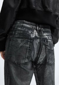 Vaqueros de mezclilla negros con un acabado brillante y texturizado. Incluyen dos bolsillos traseros, trabillas para cinturón y detalles de costura visibles.