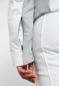 Tvåfärgad jacka med en slät grå axel och vit kropp, med en zippered ficka på vänster ärm och en figurnära design.