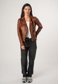 Veste de biker en cuir marron avec fermeture éclair asymétrique, finition texturée et col ajustable. Portée sur un haut blanc et associée à un jean foncé.