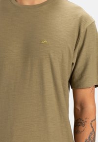 Camiseta corta de manga corta en verde oliva, con cuello redondo, tejido texturizado y un pequeño logo bordado en la esquina superior izquierda.