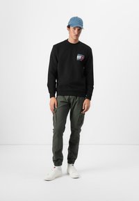 Zwarte sweatshirt met een kleurrijk logo op de borst, gecombineerd met olijfgroene cargo pants met zijzakken en witte sneakers.