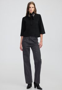 Vrouw die staat in een zwarte top met versierde kraag, donkere high-waisted jeans en zwarte enkellaarsjes met hakken, tegen een effen achtergrond.