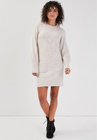 Robe pull en beige clair, dotée de manches longues, de deux poches avant et d'un ourlet côtelé. Motif en V subtil sur l'avant.