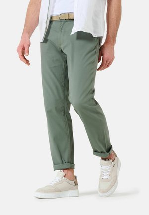 Helle grüne, schmal zulaufende Hose mit glatter Textur und umgeschlagenen Säumen, kombiniert mit beigen und weißen Sneakers, die eine strukturierte Oberseite und flache Sohle haben.