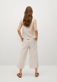 Femme aux longs cheveux châtain se tient de dos, portant un top sans manches beige, un pantalon large et court assorti, et des sandales marron.