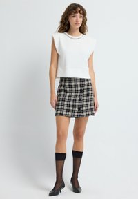 Top blanco corto con hombros estructurados y un detalle de cadena, combinado con una falda corta de cuadros en blanco y negro y calcetas negras transparentes hasta la rodilla.