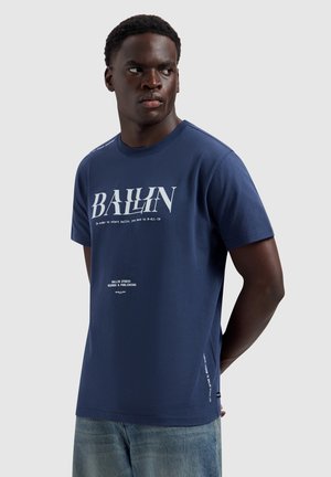Man draagt een marineblauw T-shirt met de opdruk "BALLIN" en een lichtblauwe spijkerbroek, staat met de handen achter zijn rug en kijkt naar links.