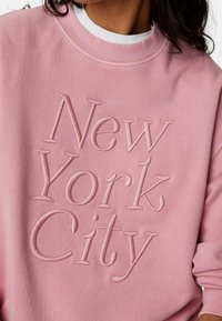 Sudadera rosa claro con texto en relieve "New York City". Cuello redondo y puños acanalados, confeccionada en una suave tela texturizada.