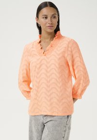 Blouse texturée couleur pêche avec un col en V à volants et des manches bouffantes trois-quarts. Présente un motif en zigzag sur l'ensemble.
