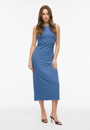 Pikkade pruunide juustega naine, kes kannab varrukateta sinist midi kleiti ja pruune kõrge kontsaga sandaale, seisab valge tausta ees.