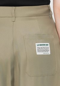 Beige stoffbukser med en baklomme som har et hvitt merke. Merket inkluderer merkevarenavnet "LA MAISON AMV" og vaskeanvisninger.