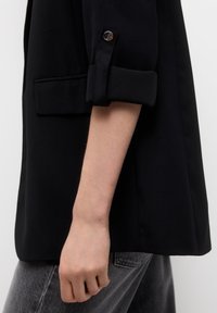 Černý blazer s ohrnutými rukávy, vyrobený z hladkého materiálu, s jediným knoflíkem na manžetě a přední kapsou. Šedé kalhoty s vysokým pasem.