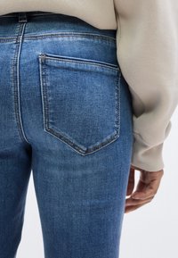 Μπλε τζιν παντελόνι με κοντινό πλάνο της πίσω τσέπης, που παρουσιάζει διακριτική ξεθωριασμένη υφή και αντίθετη ραφή, τοποθετημένο σε φωτεινό φόντο.