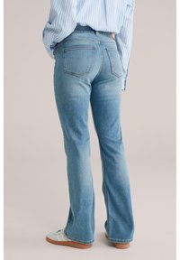 Oseba, ki nosi svetlo modre široke kavbojke, modro-belo črtasto majico in bele superge, stoji ob svetlem nevtralnem ozadju.